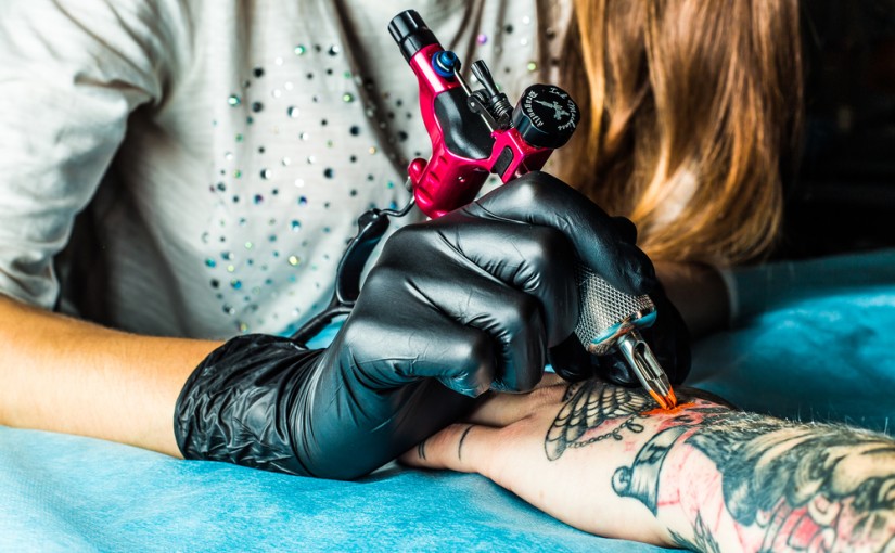 Master tattoo draws the orange paint on the clients tattoo. Tattoo artist holding a pink tattoo machine in black sterile gloves and working on the professional blue mat.